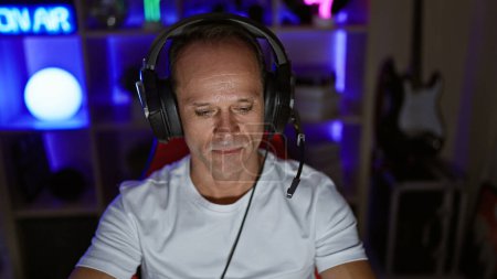 Seriöser Mann mittleren Alters, der in seinem Home Office in die digitale Welt des Spielens eintaucht - gutaussehender hispanischer Gamer, rockige Kopfhörer, die ganze Nacht Cyber-Unterhaltung streamen