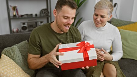 Foto de Un hombre y una mujer comparten un momento alegre en un sofá, abriendo con entusiasmo una caja de regalo roja en una acogedora sala de estar. - Imagen libre de derechos