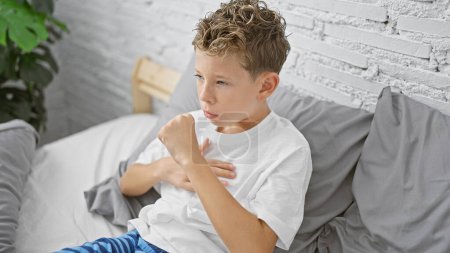 Photo for Adorable blond boy, sitting on bed in his pajamas, coughing in the early morning. sick child resting in bedroom, fighting flu, indoors in a cosy house. - Royalty Free Image