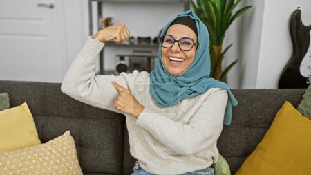 Photo for Smiling middle-aged woman flexing muscle in a cozy living room, showcasing strength and happiness. - Royalty Free Image