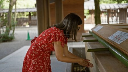 Photo for Beautiful hispanic woman with glasses participates in traditional asian purifying ritual at meiji temple, washing hands in flowing natural water from bamboo fountain for spiritual cleansing - Royalty Free Image