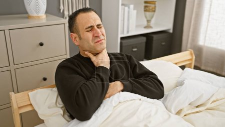 Hombre hispano en incomodidad sentado en la cama sosteniendo su cuello en un dormitorio moderno