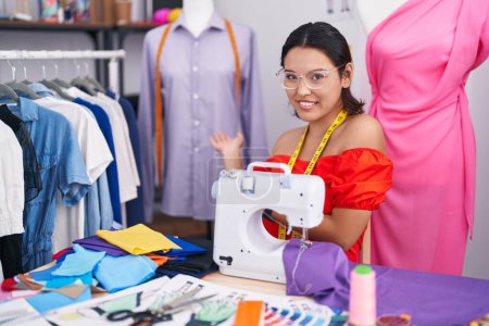 Photo for Hispanic young woman dressmaker designer using sewing machine inviting to enter smiling natural with open hand - Royalty Free Image