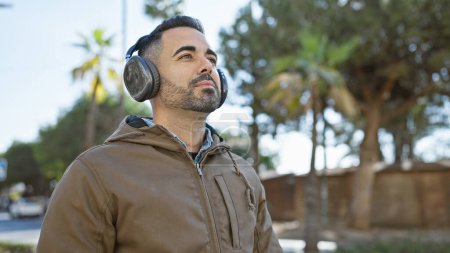 Photo for Stylish hispanic man with beard enjoying music on headphones outdoors on a sunny urban street. - Royalty Free Image