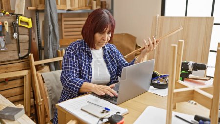 Photo for A mature hispanic woman examines a wooden part while working with a laptop in a carpentry workshop. - Royalty Free Image
