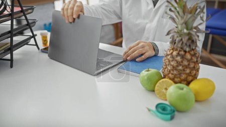 Nutricionista de capa blanca cerrando laptop en clínica con frutas y cinta métrica en escritorio