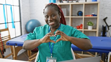 Foto de Mujer afroamericana con trenzas haciendo un gesto cardíaco en una clínica de fisioterapia. - Imagen libre de derechos
