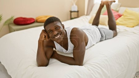 Téléchargez les photos : Homme afro-américain souriant relaxant dans un cadre confortable de chambre à coucher, dépeignant loisirs et confort à la maison. - en image libre de droit