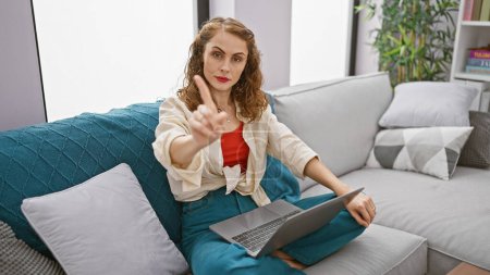 Focused young woman indoors on sofa - saying no with a finger gesture while using laptop, engrossed in serious online work at home