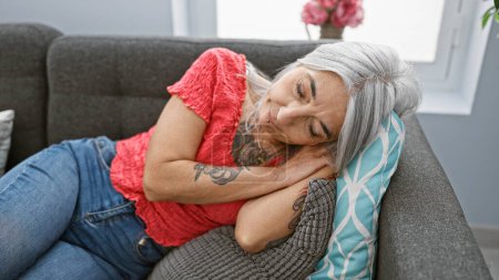 Entspannte, schöne grauhaarige Frau mittleren Alters findet Trost in Ruhe, im Liegen und Schlafen auf einem gemütlichen Sofa im Haus, das ihr Zufluchtsort ist, wo die Müdigkeit nachlässt