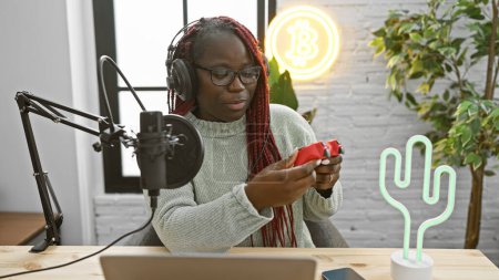 Femme afro-américaine avec des tresses examine un joystick dans un studio de radio intérieur moderne.