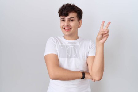 Téléchargez les photos : Jeune homme non binaire portant un t-shirt blanc décontracté souriant avec un visage heureux clin d'oeil à la caméra faisant signe de victoire avec les doigts. numéro deux. - en image libre de droit