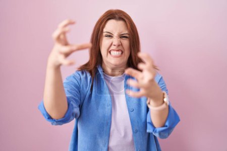 Photo for Young hispanic woman with red hair standing over pink background shouting frustrated with rage, hands trying to strangle, yelling mad - Royalty Free Image