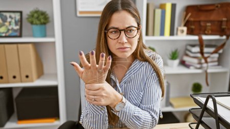 Téléchargez les photos : Une femme soucieuse de lunettes examine son poignet dans un cadre de bureau moderne, mettant en valeur les soins de santé sur le lieu de travail. - en image libre de droit