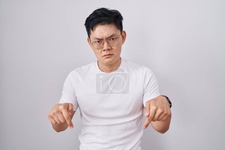 Junger asiatischer Mann steht über weißem Hintergrund und zeigt traurig und aufgebracht nach unten, zeigt mit den Fingern Richtung an, unglücklich und deprimiert. 