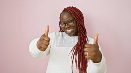 Une femme africaine joyeuse avec des tresses portant des lunettes sur un fond rose donnant pouces vers le haut.