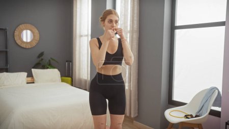 Foto de Mujer atlética en ropa deportiva bebiendo agua en un dormitorio moderno después de hacer ejercicio, lo que refleja un estilo de vida saludable. - Imagen libre de derechos