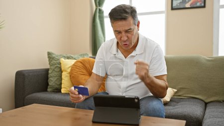 Téléchargez les photos : Homme mûr dans le salon avec tablette et carte de crédit montre surprise lors de vos achats en ligne - en image libre de droit