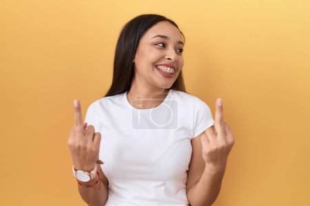 Foto de Young arab woman wearing casual white t shirt over yellow background showing middle finger doing fuck you bad expression, provocation and rude attitude. screaming excited - Imagen libre de derechos