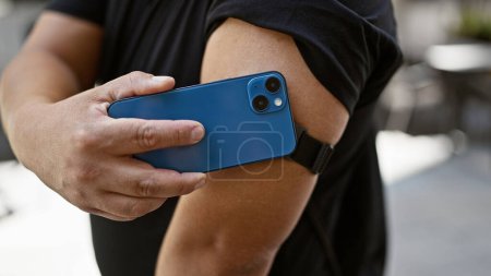 Téléchargez les photos : Jeune homme latin confiant exploitant la technologie, scannant son capteur de diabète avec un smartphone dans la rue de la ville, surveillant les niveaux de glucose avec un sourire - en image libre de droit