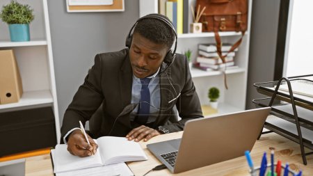 Foto de Un hombre afroamericano enfocado usando un auricular mientras escribe notas en una oficina. - Imagen libre de derechos