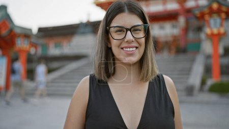 Beautiful hispanic woman with glasses poses at fushimi inari-taisha, joy radiating from her successful smile, confidence stands strikingly in kyoto's grand shrine