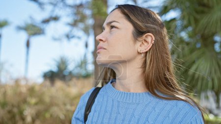 Foto de Una mujer reflexiva con ojos azules y cabello largo y morena mira serenamente en un entorno soleado y verde del parque. - Imagen libre de derechos
