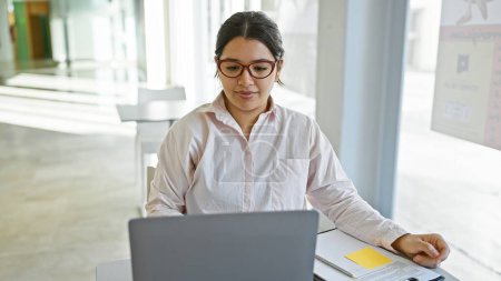 Foto de Una mujer hispana enfocada trabaja en su computadora portátil en una oficina moderna y brillante, ejemplificando profesionalismo y diligencia. - Imagen libre de derechos