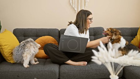 A young hispanic woman interacts cheerfully with her pet dogs while seated on a couch indoors with a laptop.