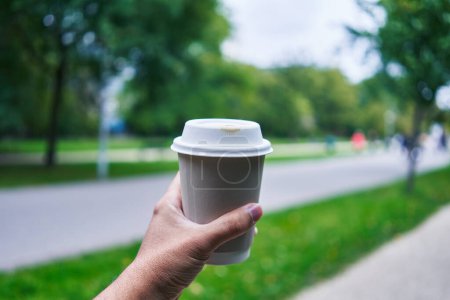 Téléchargez les photos : Un homme tient une tasse de café à emporter à l'extérieur dans un parc flou, symbolisant le mode de vie urbain et le rafraîchissement. - en image libre de droit