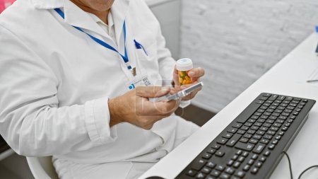 Photo for A mature man in a lab coat examines medication in a hospital setting with a smartphone and keyboard nearby. - Royalty Free Image