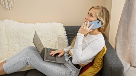 Téléchargez les photos : Une jeune femme blonde à la maison communique sur un smartphone tout en utilisant son ordinateur portable, respirant confort et connectivité. - en image libre de droit