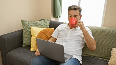 Mature man in a casual home setting drinking coffee while working on a laptop, depicting a relaxed telecommuting scene.