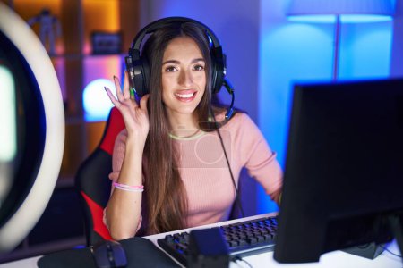 Foto de Mujer hispana joven jugando videojuegos sonriendo con la mano sobre el oído escuchando un rumor o chismes. concepto de sordera. - Imagen libre de derechos