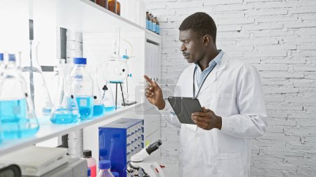 Foto de Un científico africano con una bata de laboratorio usa una tableta mientras examina el equipo en un laboratorio moderno. - Imagen libre de derechos