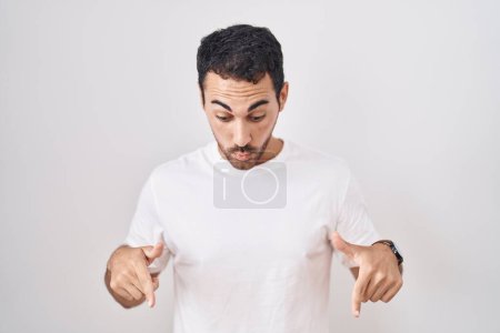 Foto de Hombre hispano guapo de pie sobre fondo blanco apuntando hacia abajo con los dedos mostrando publicidad, cara sorprendida y boca abierta - Imagen libre de derechos