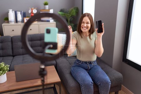 Photo for Beautiful brunette woman recording vlog tutorial with smartphone at home screaming proud, celebrating victory and success very excited with raised arm - Royalty Free Image