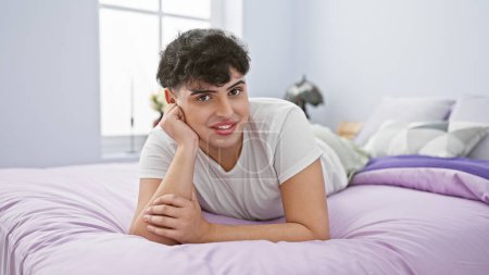 Téléchargez les photos : Jeune homme souriant couché sur un lit violet dans une chambre lumineuse, dépeignant détente et confort à la maison. - en image libre de droit