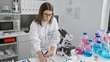 Una mujer hispana enfocada trabajando diligentemente en un laboratorio rodeado de equipos científicos y vasos de precipitados.