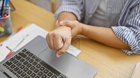 Ein Mann in einem Büro zuckt vor Schmerzen zusammen und klammert sein Handgelenk über einen Laptop, was auf ein Karpaltunnelsyndrom oder eine Verletzung hindeutet..