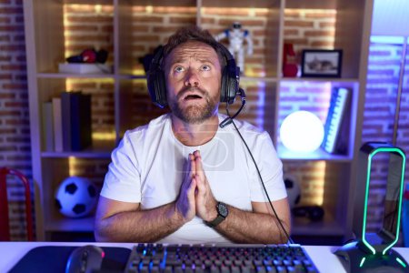 Middle age man with beard playing video games wearing headphones begging and praying with hands together with hope expression on face very emotional and worried. begging. 