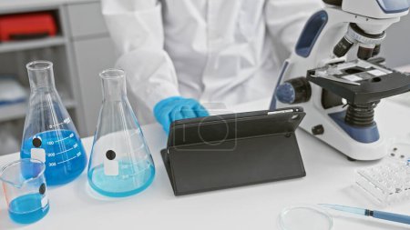 Foto de Una mujer científica utiliza una tableta en un laboratorio con un microscopio y vasos de precipitados, que representan la investigación y la tecnología en el interior. - Imagen libre de derechos