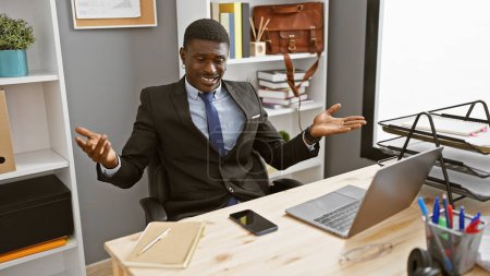 Téléchargez les photos : Homme d'affaires africain confiant gesticulant assis à son bureau avec ordinateur portable et smartphone dans un bureau moderne. - en image libre de droit