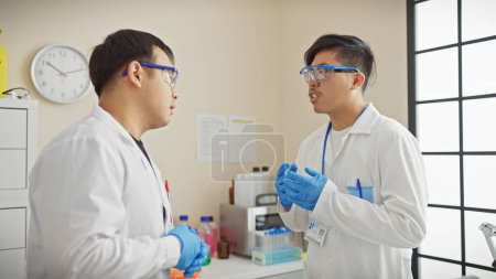 Two male scientists in discussion inside a brightly lit laboratory, wearing lab coats and protective goggles.