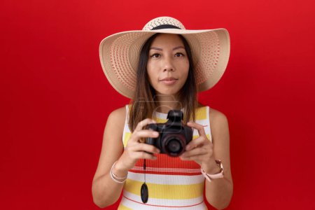 Middle age chinese woman using reflex camera wearing summer hat relaxed with serious expression on face. simple and natural looking at the camera. 