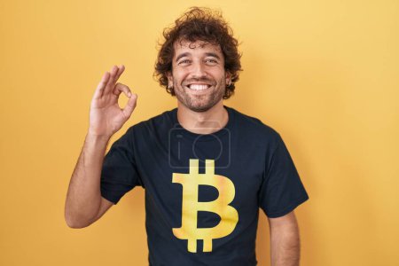 Téléchargez les photos : Jeune homme hispanique portant Bitcoin t-shirt souriant positif faire ok signe avec la main et les doigts. expression réussie. - en image libre de droit