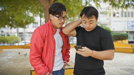 Photo for Two asian men using a smartphone together in an urban park setting. - Royalty Free Image