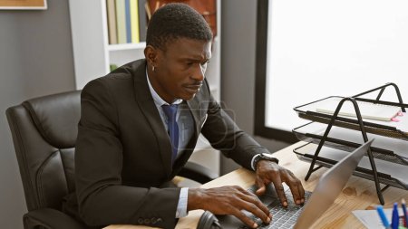 Foto de Un hombre de negocios afroamericano enfocado en un traje que trabaja en su escritorio de oficina con una computadora portátil y monitor. - Imagen libre de derechos