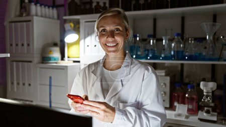 Sonriente mujer caucásica en bata de laboratorio usando smartphone en un interior de laboratorio.