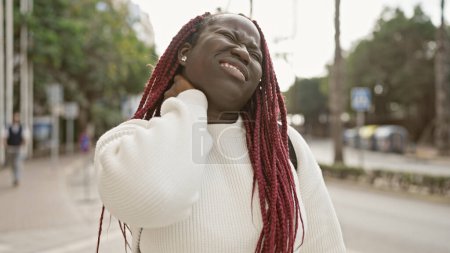 Téléchargez les photos : Femme afro-américaine avec des tresses exprimant une douleur au cou à l'extérieur dans un cadre urbain. - en image libre de droit